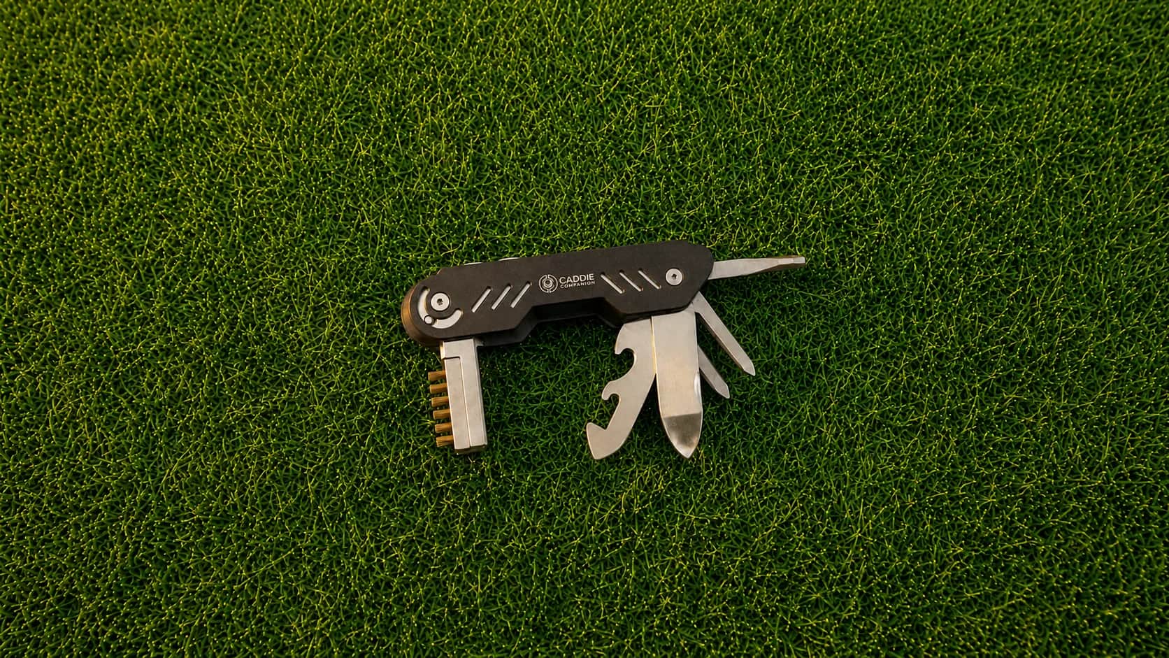 Black Caddie Companion golf multi-tool open on the grass, showing its divot fork, knife, and groove brush