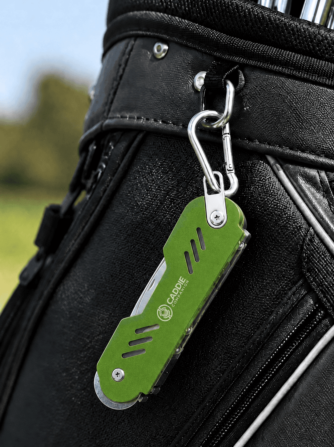 Green Caddie Companion golf tool clipped by its carabiner to the side of a black golf bag on the course